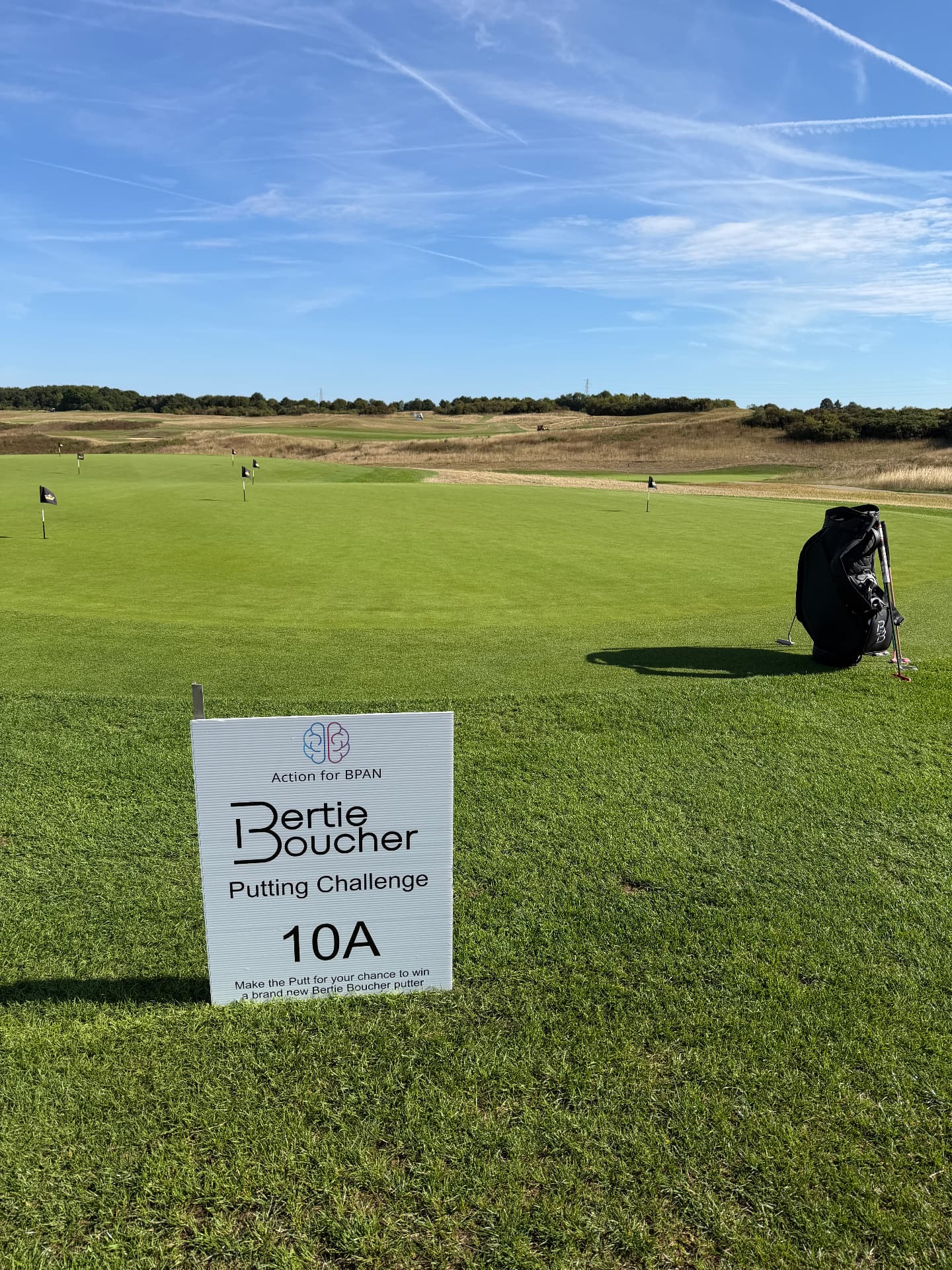 Action for BPAN Bertie Boucher Putting Challenge sign on the putting green with hole flags and a golf bag